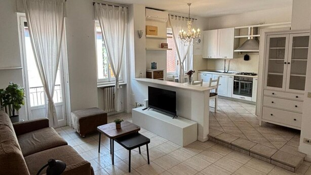 Una sala de estar moderna con un sofá, televisión y una mesita. Hay una zona de cocina con armarios y una mesa de comedor.