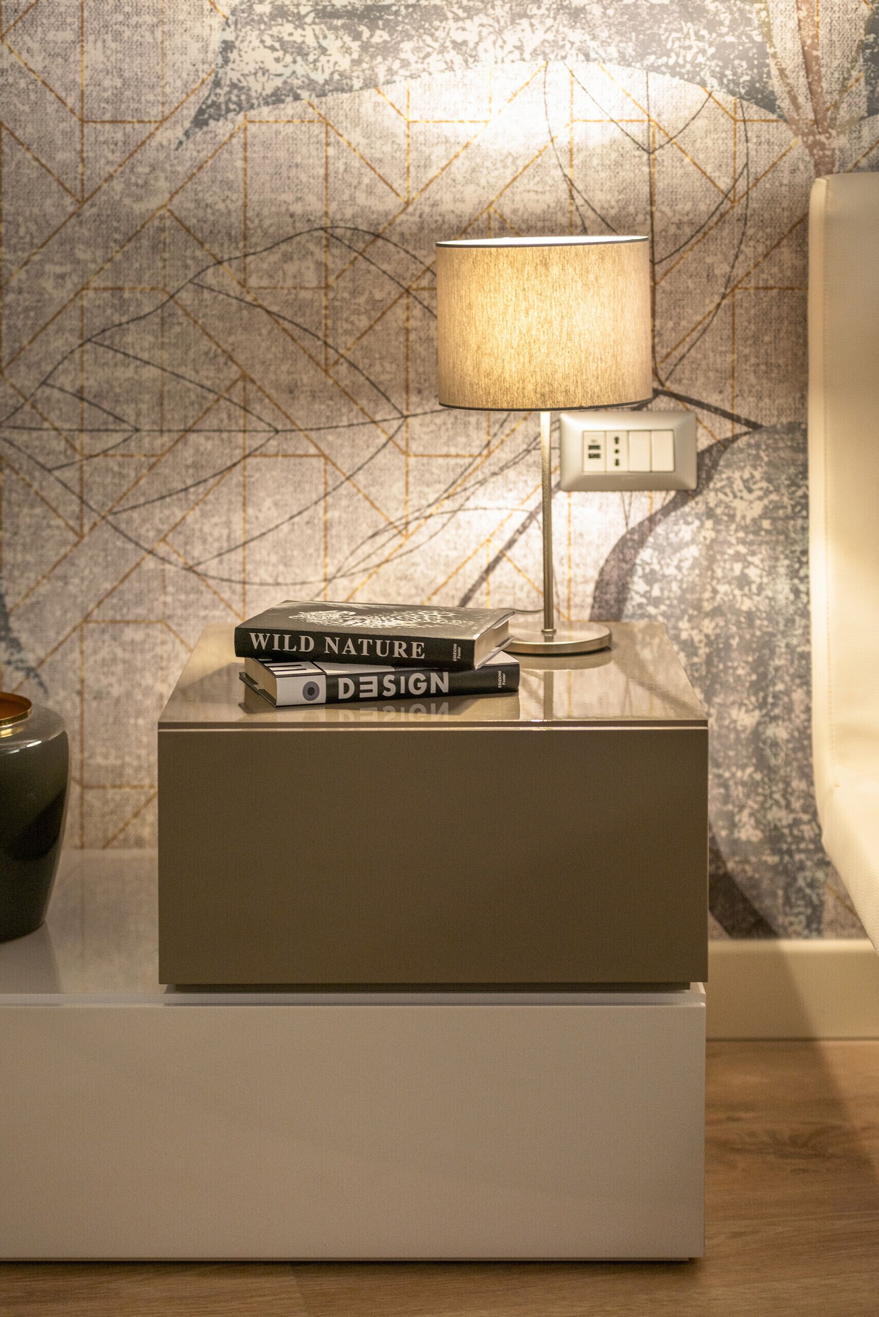 A bedside table with a lamp, two books titled 'Wild Nature' and 'Design', and a power outlet.