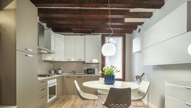 Una cocina moderna con vigas de madera, armarios blancos, una mesa redonda y un jarrón azul con flores.