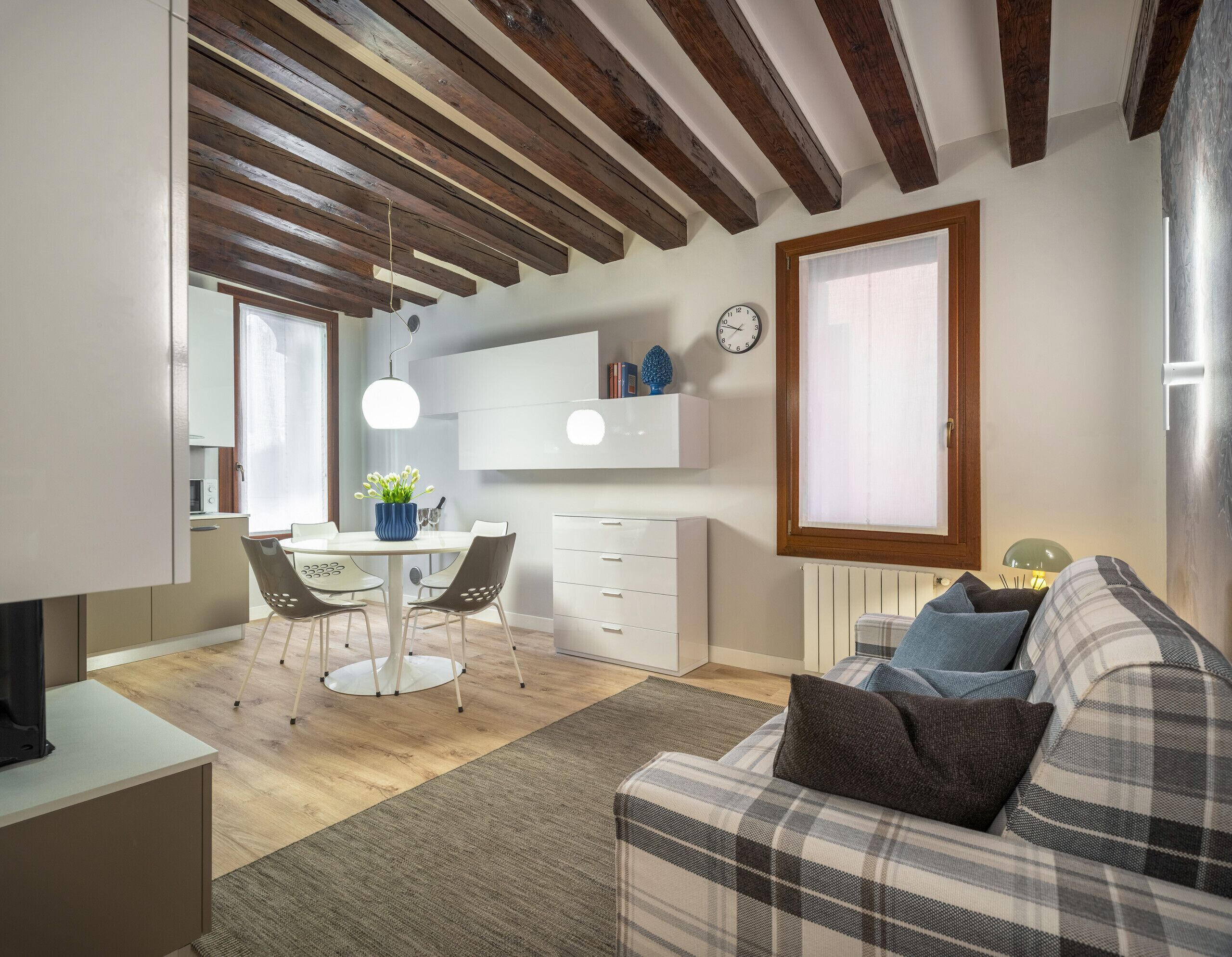 Ein modernes Wohnzimmer mit einem Karomuster-Sofa, einem Essbereich mit weißem Tisch und Stühlen sowie einer Küche mit weißen Schränken und einer Uhr an der Wand.