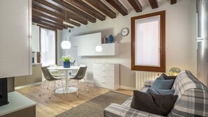 Una sala de estar moderna con un sofá a cuadros, un comedor con una mesa y sillas blancas, y una cocina con armarios blancos y un reloj en la pared.