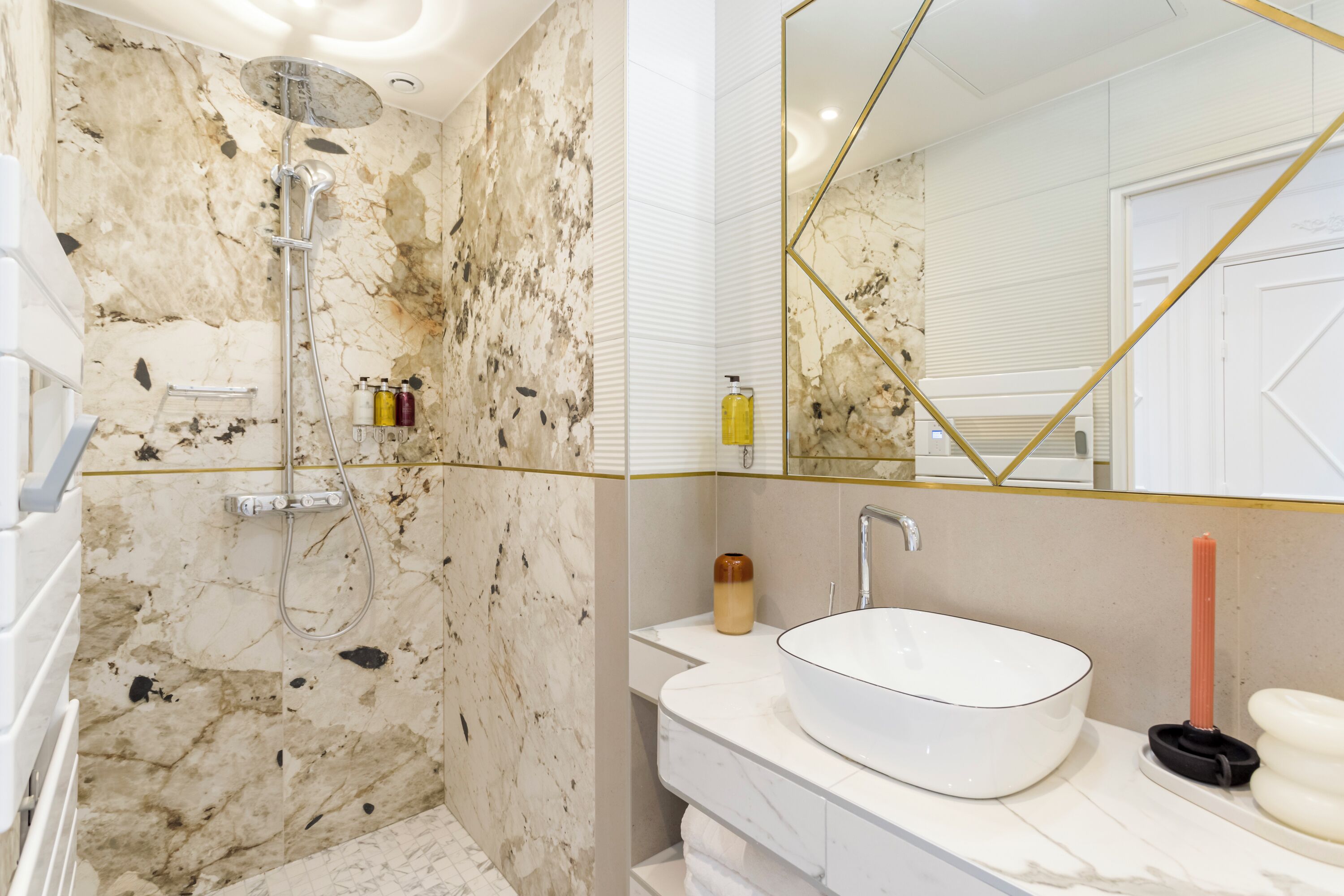 A bathroom with a marble shower, a white sink, and a large mirror with a geometric gold frame.