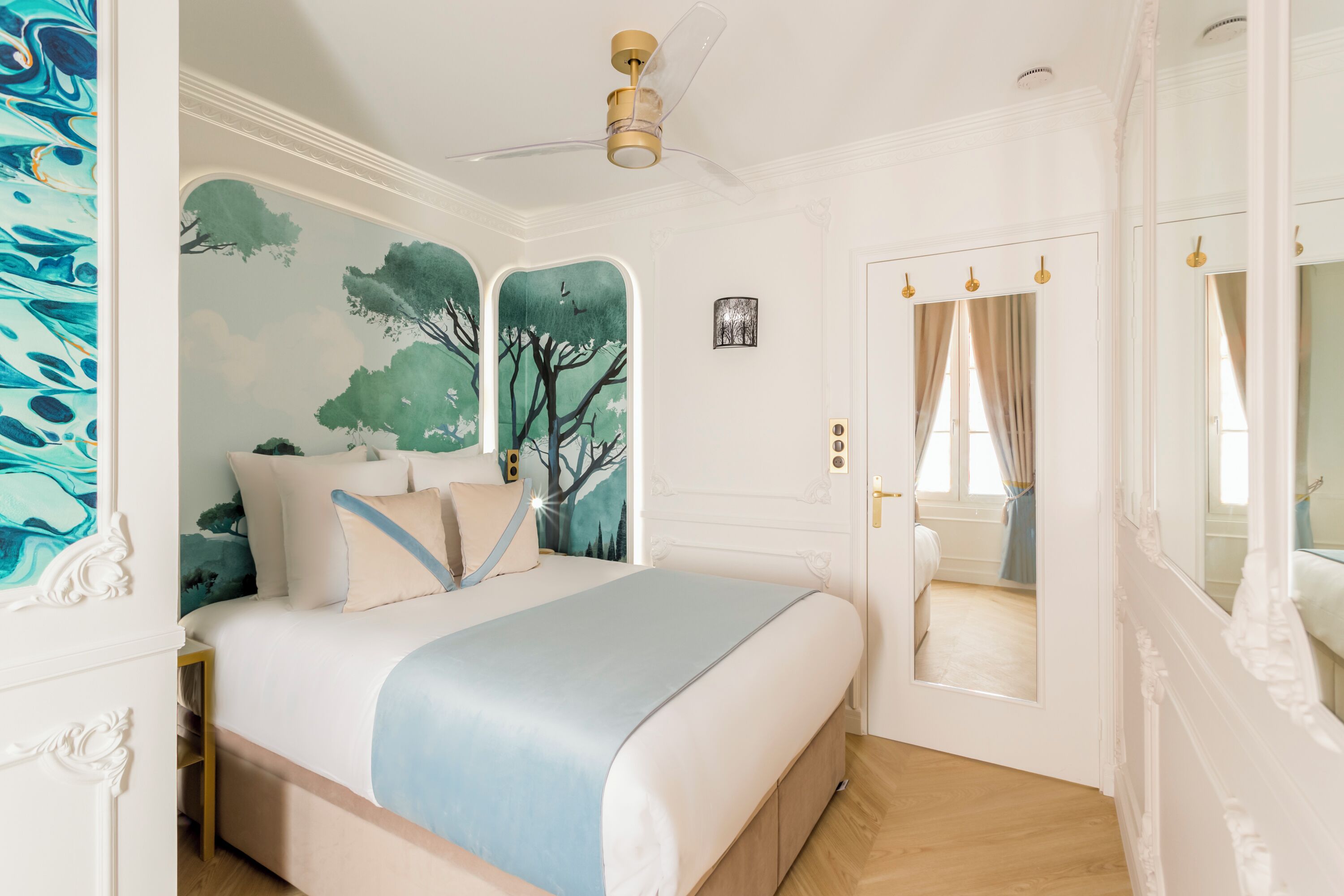 A bedroom with a bed, a ceiling fan, a door with a mirror, and a wall with a tree mural.