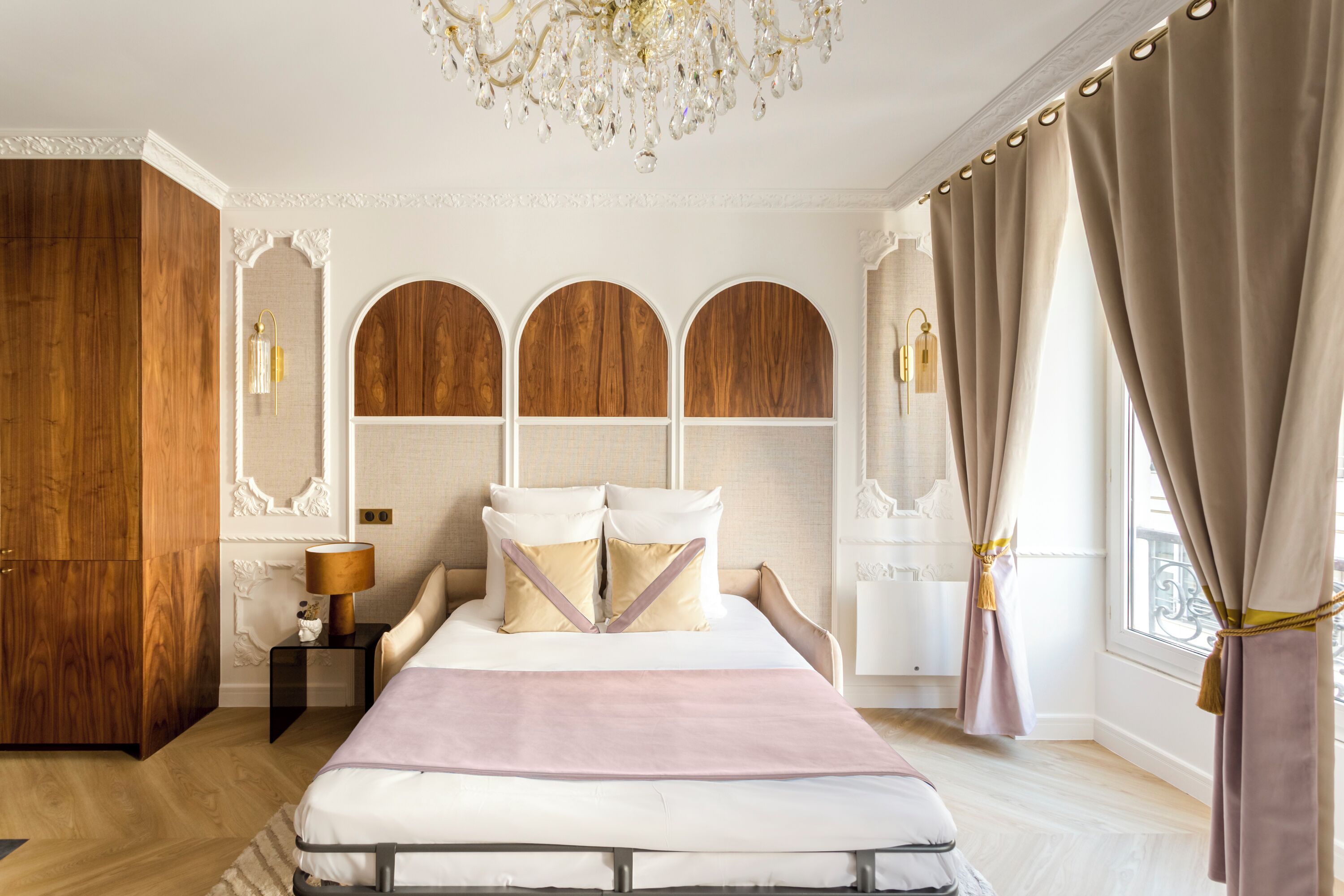 A bedroom with a large bed, wooden paneling, a chandelier, and a view through curtains.