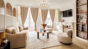 En rummelig stue med en beige sofa, et rundt sofabord og en hvid lænestol. En lysekrone hænger fra loftet, og et fladskærms-tv er monteret på væggen.