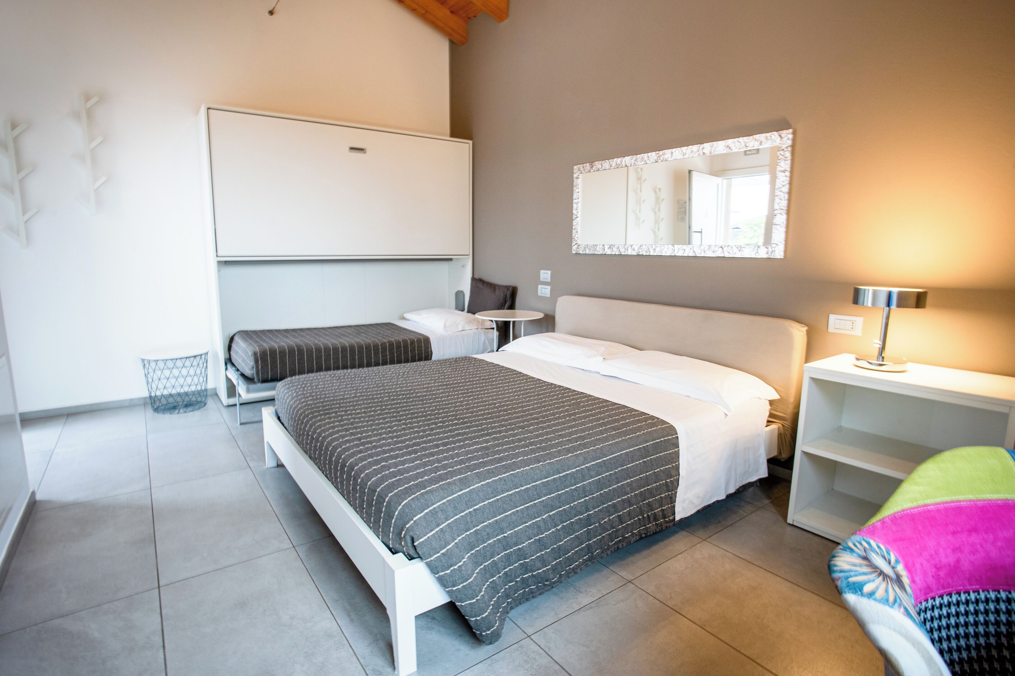 A hotel room with a large bed, a smaller bed, a bedside table, a nightstand, a chair, a lamp, a mirror, and a wall-mounted shelf.