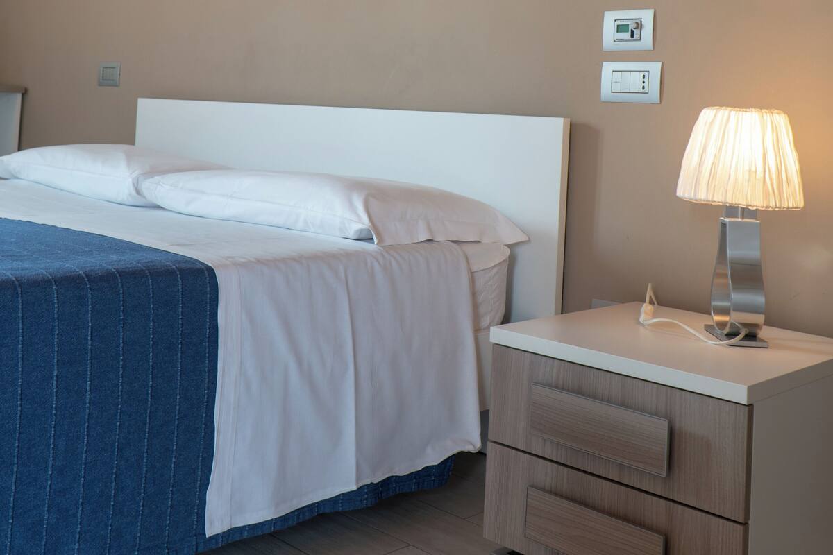 A hotel room with a bed, a nightstand with a lamp, and two wall-mounted light switches.
