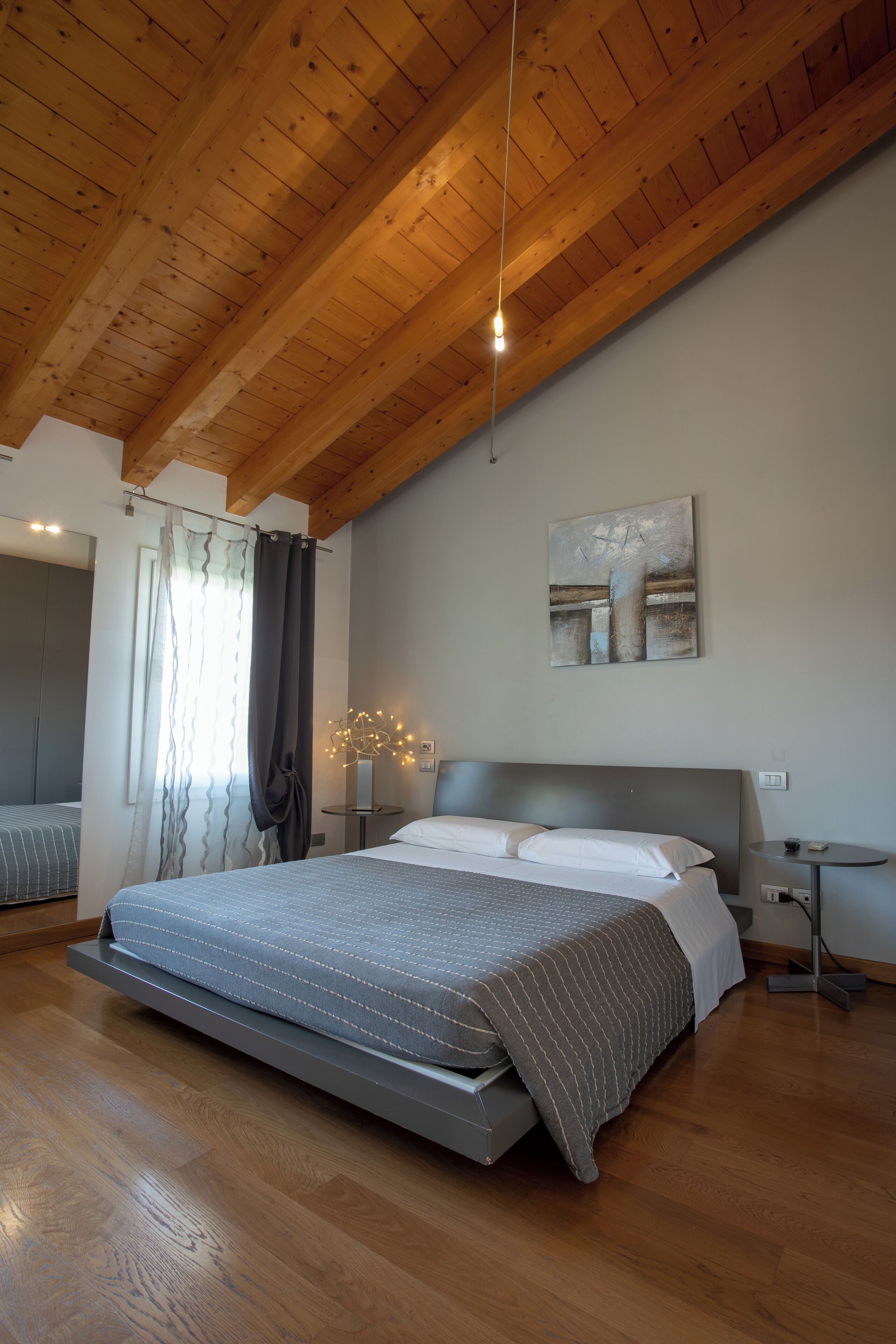 A bedroom with a wooden ceiling, a bed with a grey and white striped bedspread, a bedside table with a lamp, and a wall-mounted abstract painting.