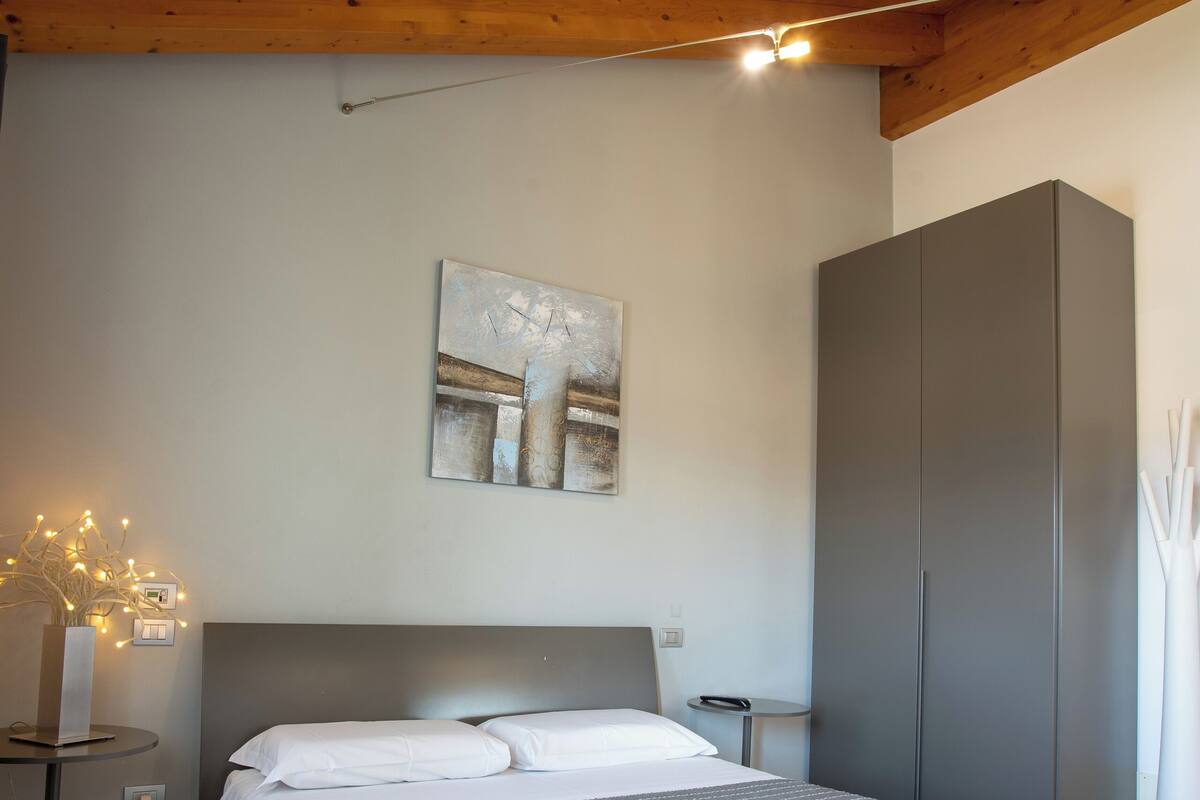 A modern bedroom with a wooden ceiling, a bed with a grey striped bedspread, a grey wardrobe, and a wall-mounted abstract painting.