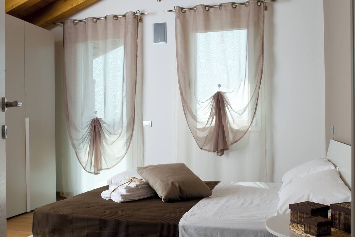 A bedroom with a loft bed, a bedside table, and sheer curtains.