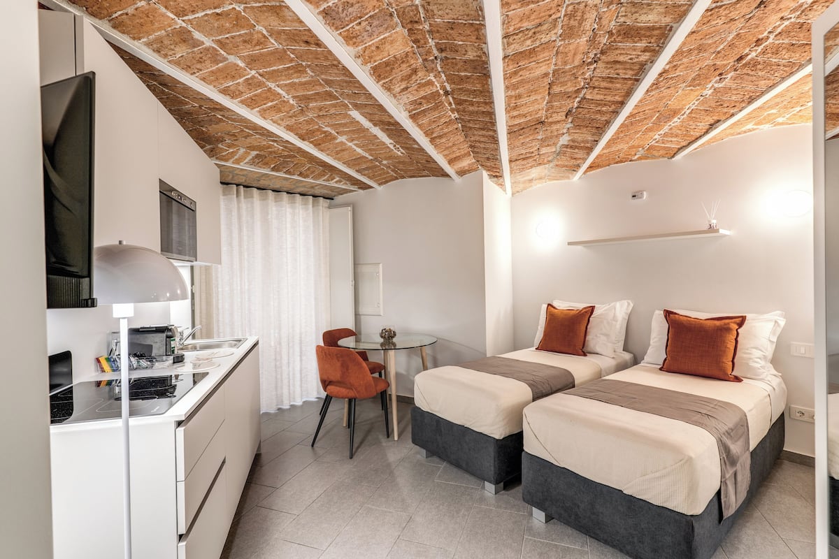 A modern room with a brick ceiling, a kitchenette, two beds with brown pillows, a small table, and a TV.