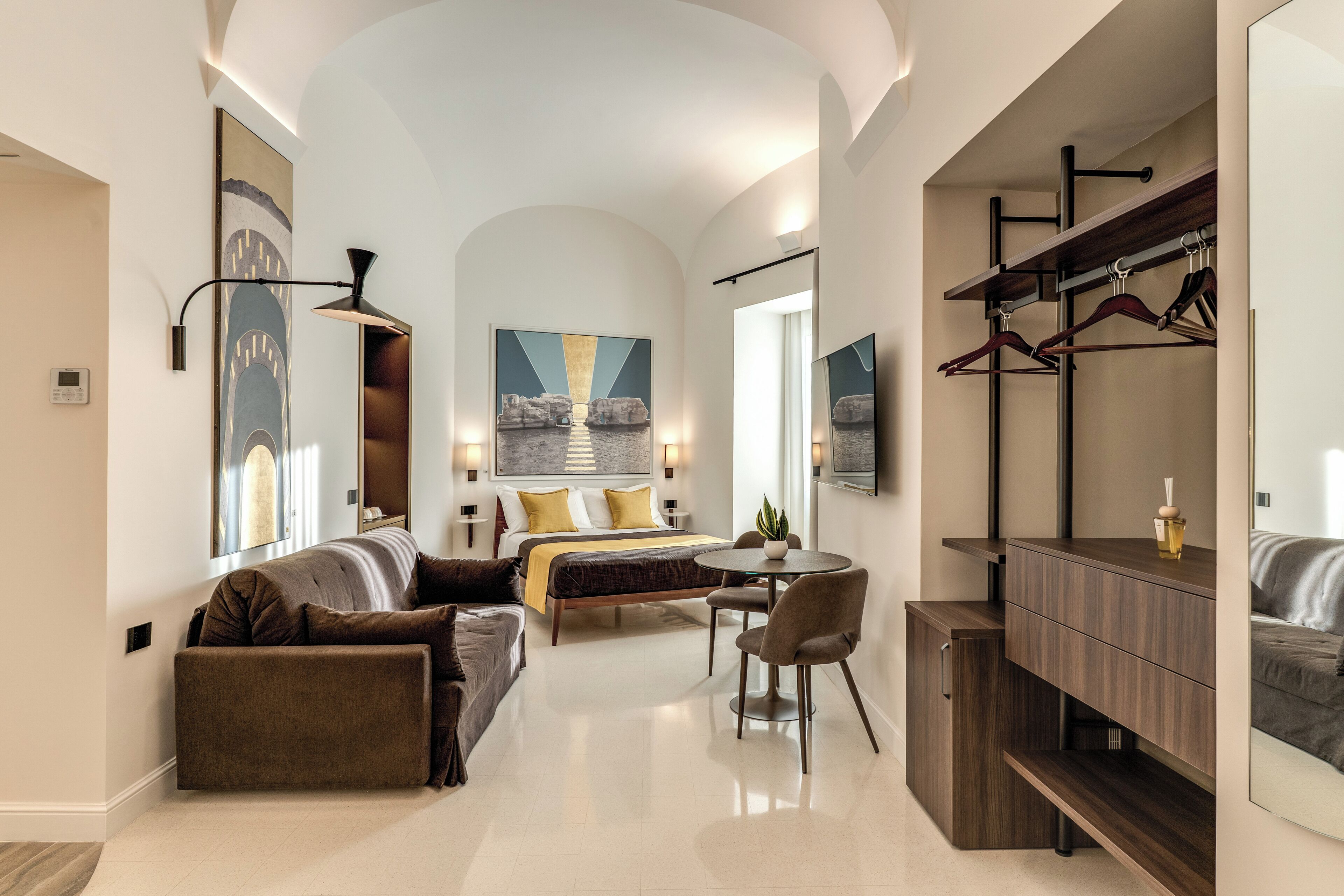 A modern hotel room with a bed, a sofa, a dining table, and a mirror.
