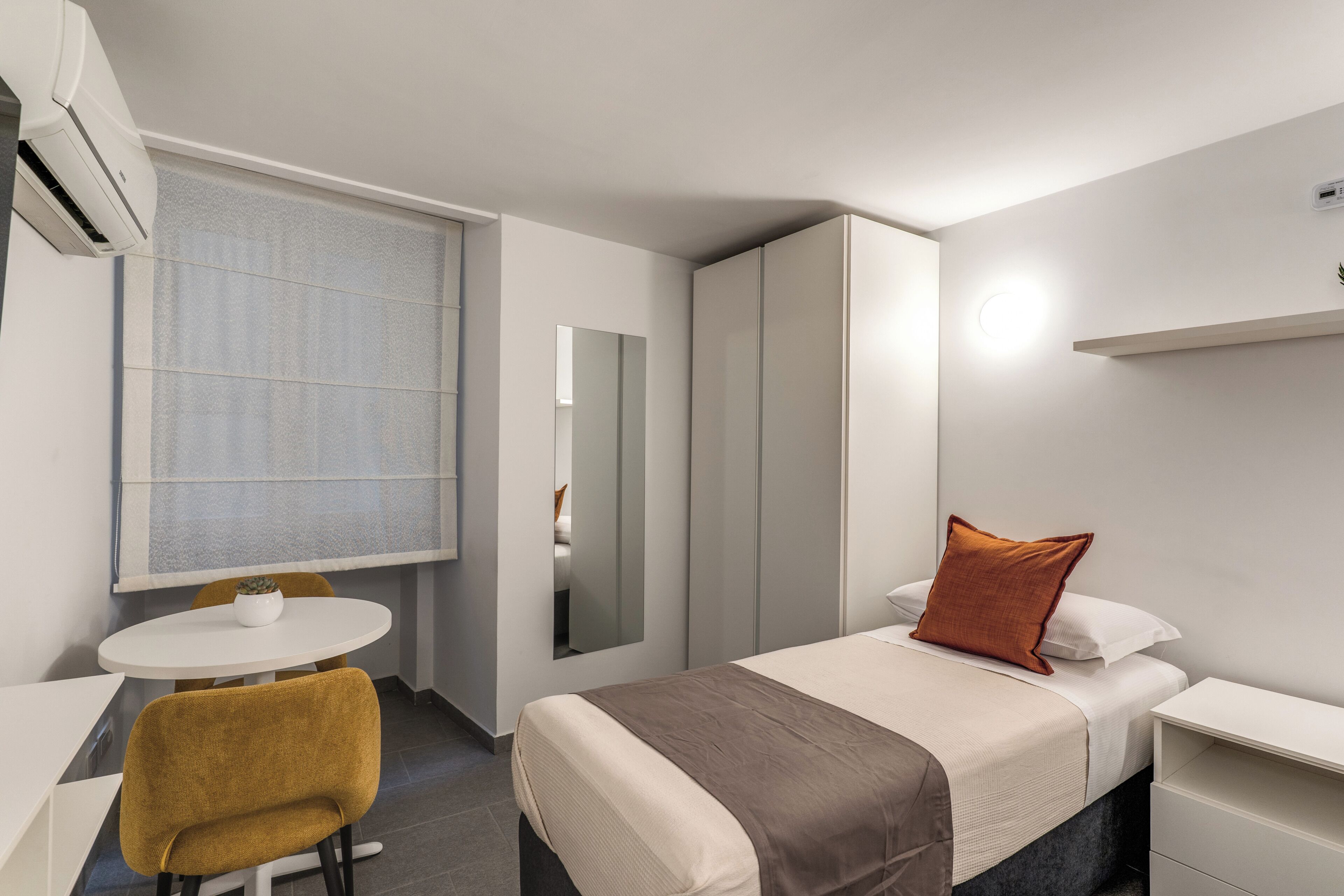 A modern hotel room with a bed, a desk, a chair, a window, and an air conditioning unit.