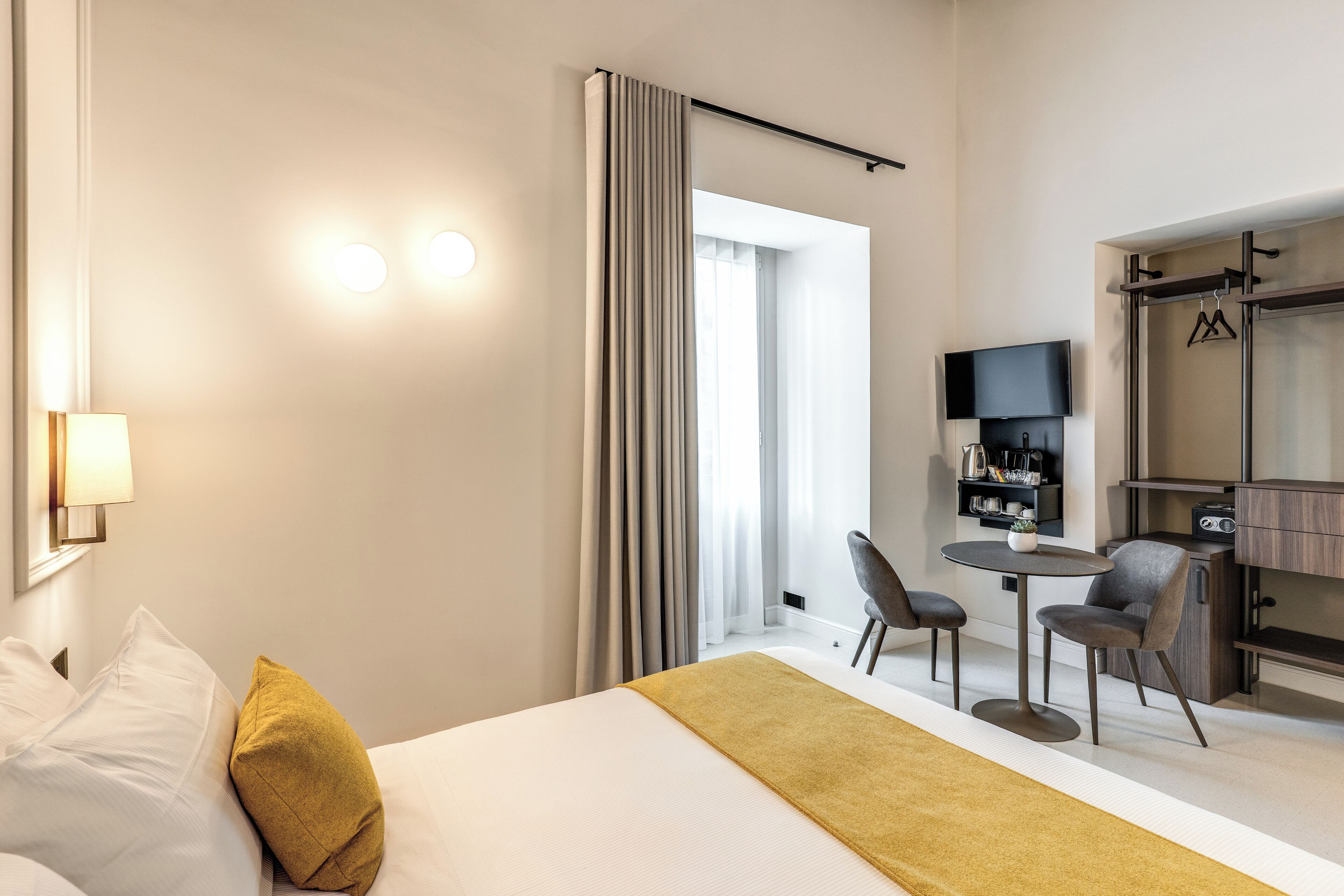 A modern hotel room with a bed, a desk, two chairs, a TV, and a wardrobe.