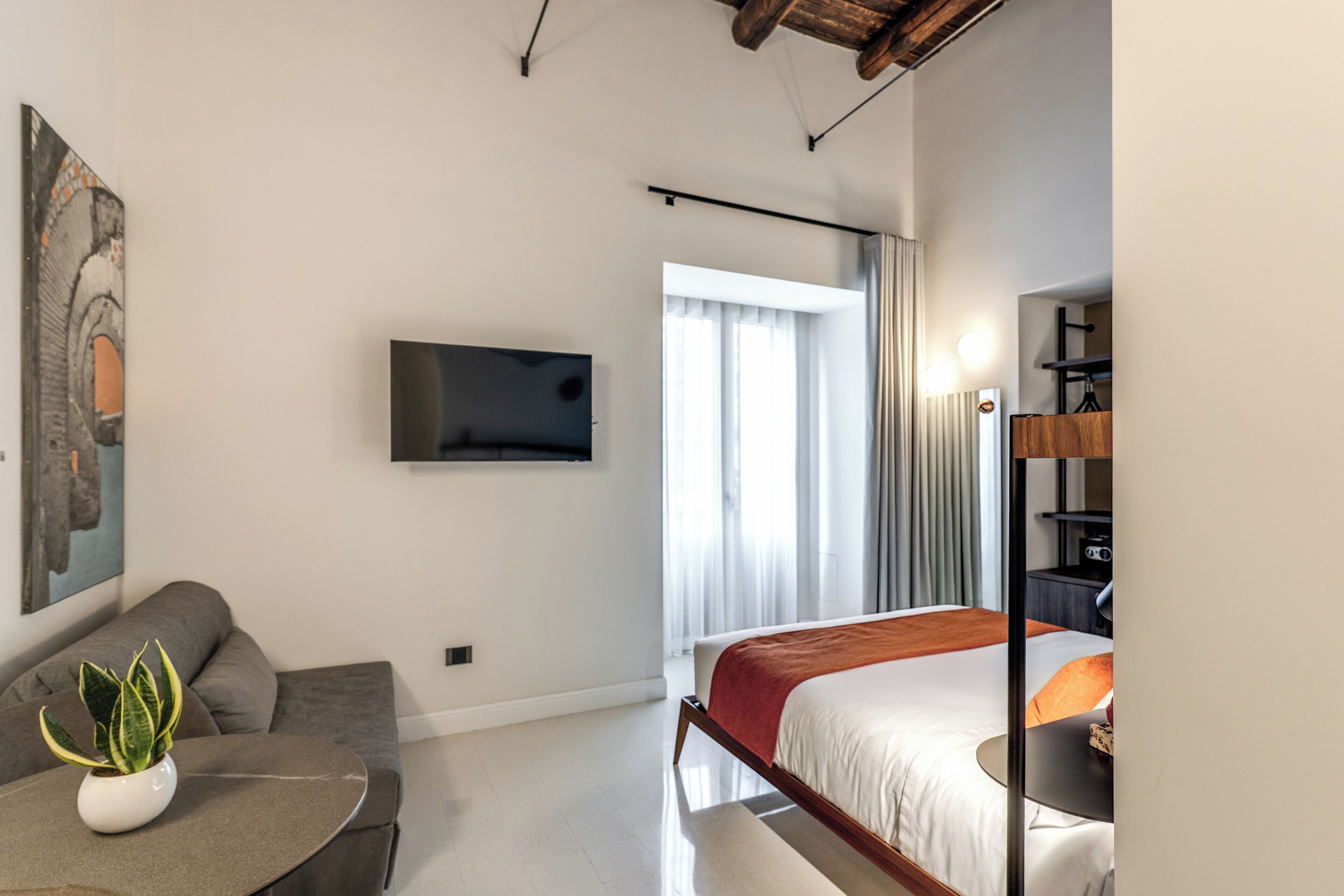 A modern hotel room with a bed, a seating area with a plant, a flat-screen TV mounted on the wall, and a window with curtains.
