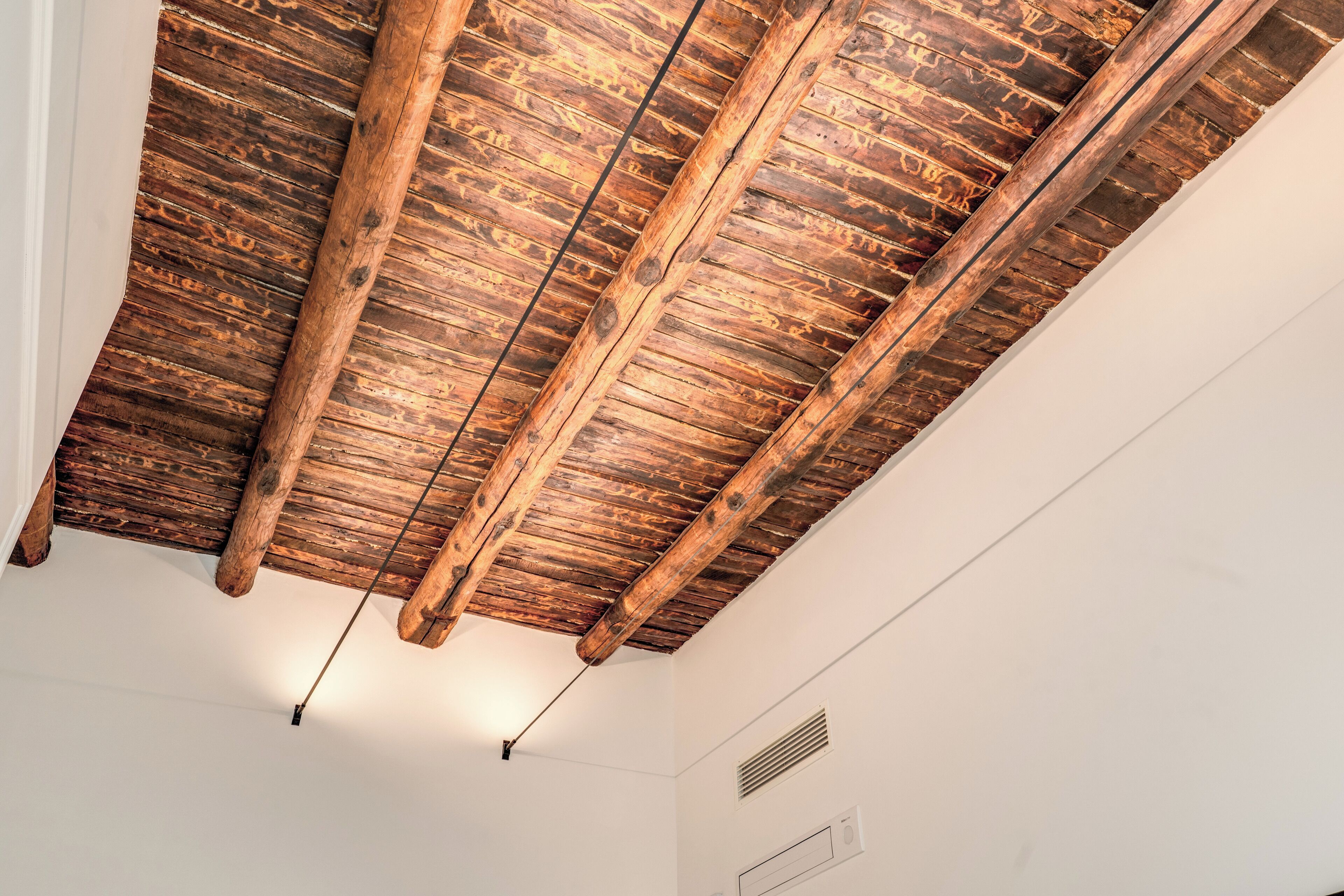 A ceiling with exposed wooden beams and a white wall with a vent.