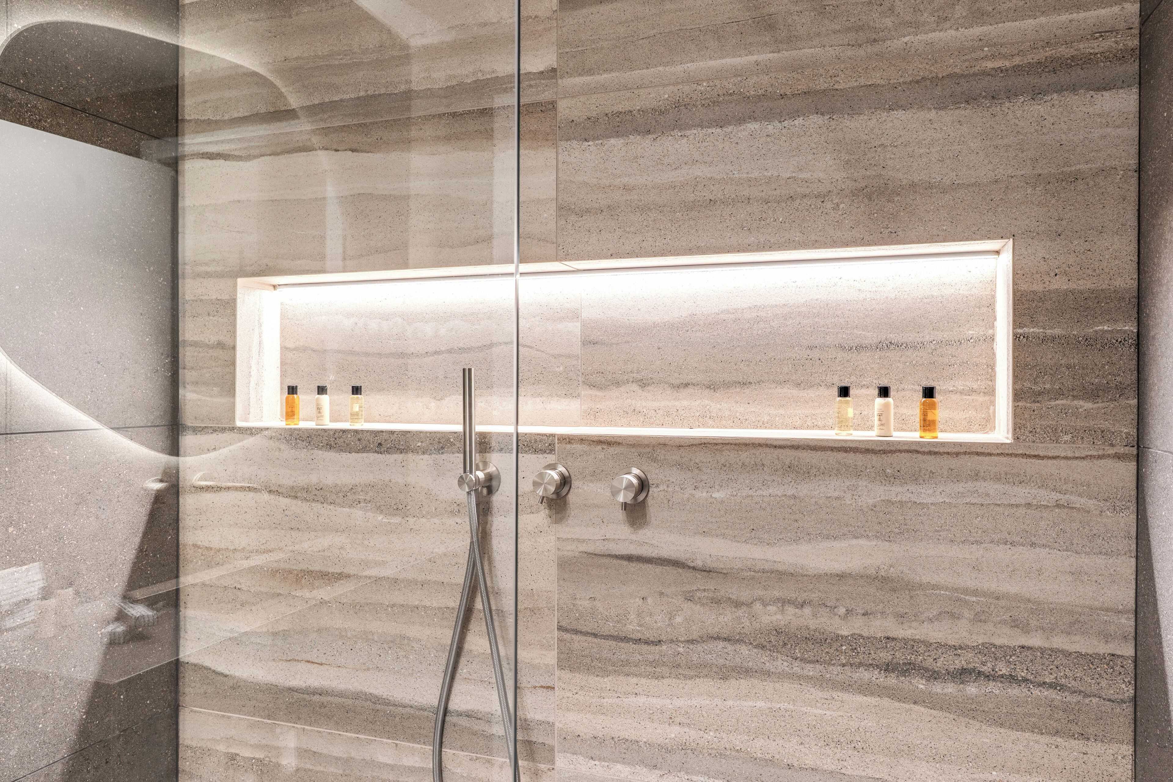 A modern bathroom with a glass shower enclosure, a marble wall, and a shelf with toiletries.