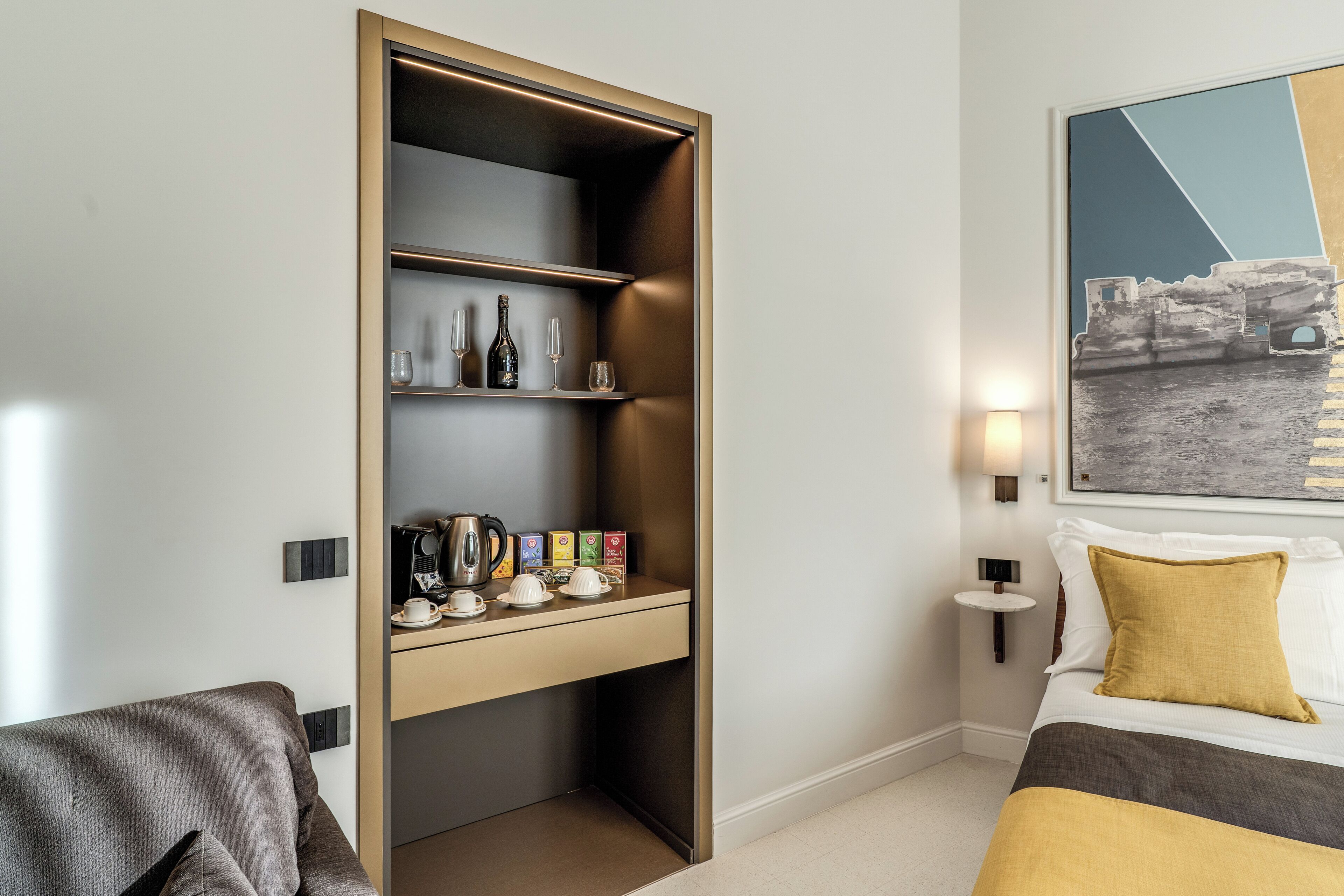 Een hotelkamer met een bed, een sofa, een inbouwplank met flessen en glazen, een kleine tafel met kopjes en een waterkoker, en een wandlamp.
