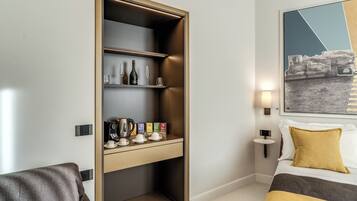 Een hotelkamer met een bed, een sofa, een inbouwplank met flessen en glazen, een kleine tafel met kopjes en een waterkoker, en een wandlamp.