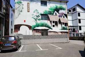 Un bâtiment orné d  ’une fresque représentant une maison et des arbres, un parking et une voiture.