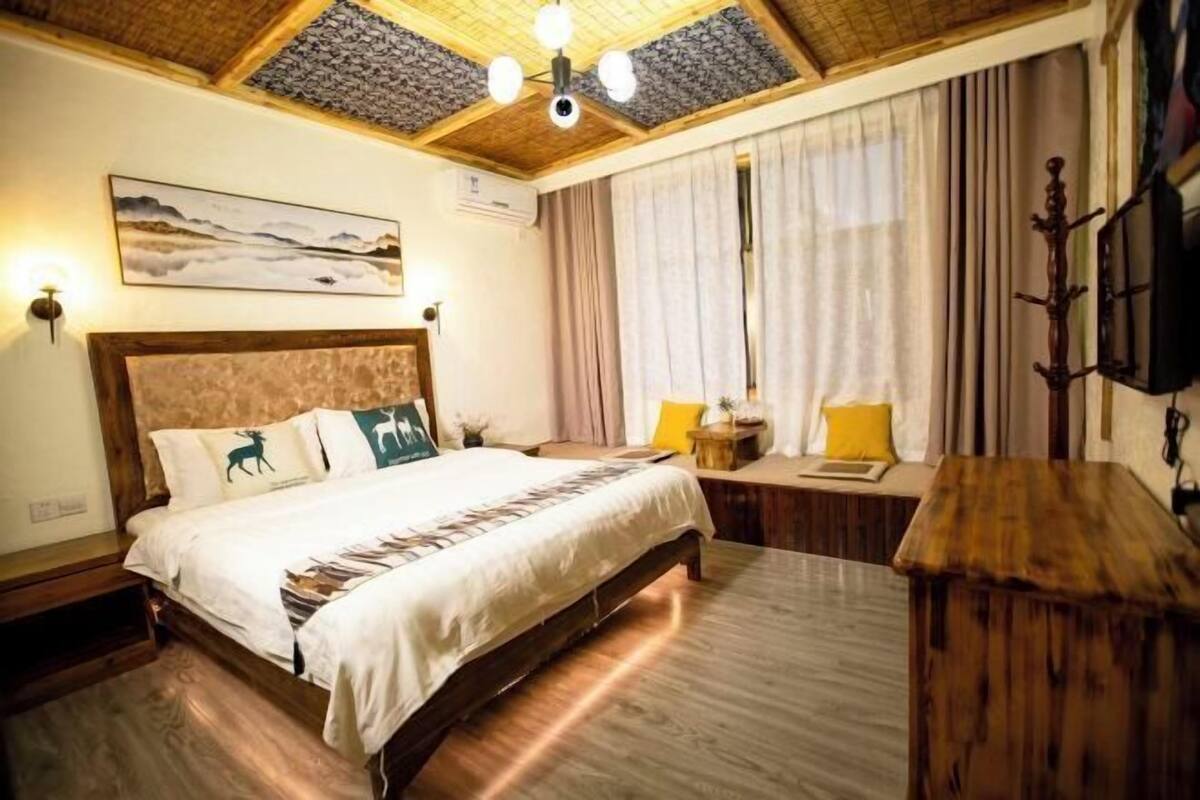 Een slaapkamer met een houten bed, een nachtkastje, een lamp, een raam met gordijnen en een aan de muur bevestigde televisie.