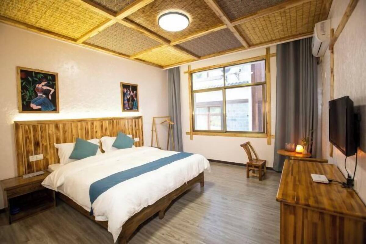 Hotelkamer met een bed, houten hoofdeinde, een bureau, een stoel, een televisie, een raam en een plafondventilator.