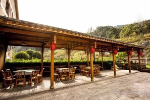 Un espace de restauration en plein air, avec des tables et des chaises en bois, abrité par une pergola décorée de lanternes rouges, le tout se fondant dans un décor de verdure et de collines.