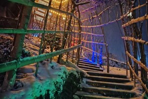Un escalier en bois avec une rampe en métal, entouré de neige et éclairé par des lumières chaleureuses.