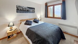 Une chambre à coucher comprenant un lit, des tables de chevet, un radiateur, une fenêtre avec des rideaux et une œuvre d’art murale.