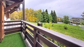 Un balcon en bois offrant une vue sur une pelouse verdoyante, des arbres et une ville lointaine.