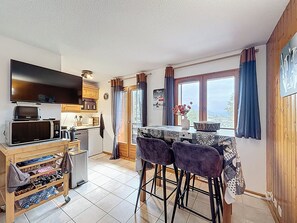 Een kleine keuken met een magnetron, broodrooster en een kleine koelkast. Er is een eethoek met een tafel en vier stoelen. De keuken heeft een raam met gordijnen en uitzicht op de omgeving.