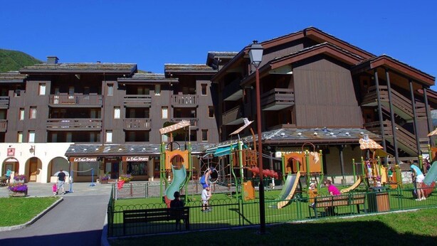 A multi-story hotel with balconies, a children's playground, and a ski resort in the background.