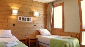 A room with two beds, wooden walls, a window with curtains, and wall-mounted lights.