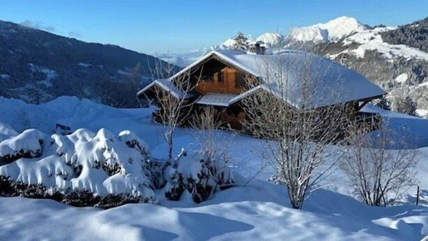 Una baita di legno con tetto spiovente, circondata da montagne e alberi innevati.