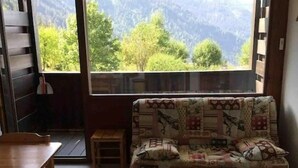 Una camera accogliente con una panca in legno, un tavolino e un divano decorato. Da una grande finestra si apre una vista su alberi verdi e montagne.