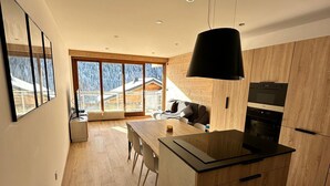 A modern kitchen with wooden cabinets, a central island, and a large window offering a view of a house. - 2-room apartment in Châtel, sleeps 5 with WIFI and pets welcome (Châtel)