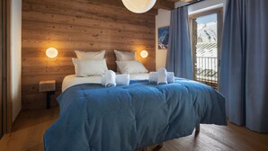 A bedroom with a wooden ceiling, a large bed with blue bedding, two bedside lamps, and a window with curtains. - Apartment for 10 people with sauna in Saint-Martin-de-Belleville (Saint-Martin-de-Belleville)