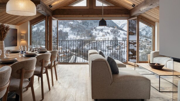 A spacious room with a high ceiling, wooden beams, a dining area with chairs and a table, a sofa with cushions, and large windows offering a view of a snowy mountain landscape. - Apartment for 10 people with sauna in Saint-Martin-de-Belleville (Saint-Martin-de-Belleville)