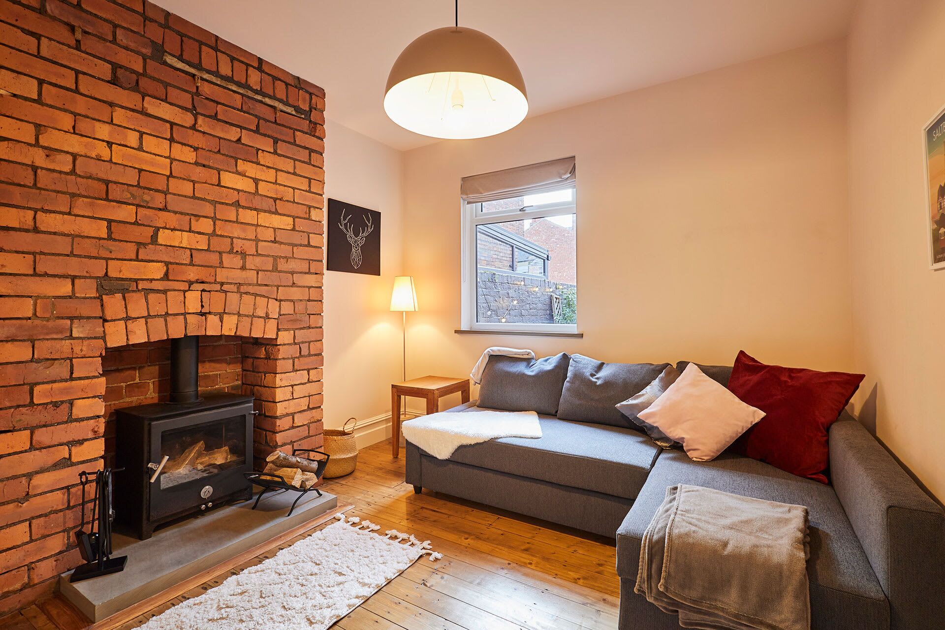 A cozy living room with a brick fireplace, a grey sofa, and a window with a view of the outside.