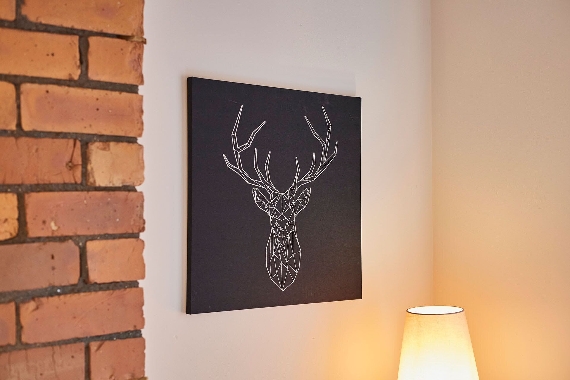 Una obra de arte con cabeza de ciervo montada en la pared en una habitación con pared de ladrillo y una lámpara encendida.