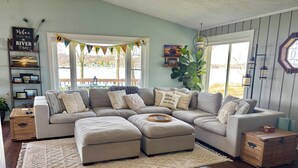 A cozy living room with a sectional sofa, ottomans, a rug, wooden side tables, a bookshelf, and a large window with a view.