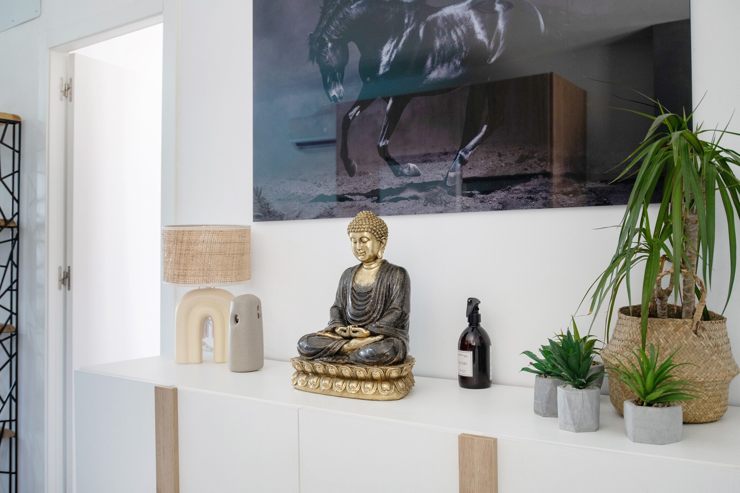 A room with a white console table, a Buddha statue, a framed horse painting, a lamp, a bottle, and potted plants.