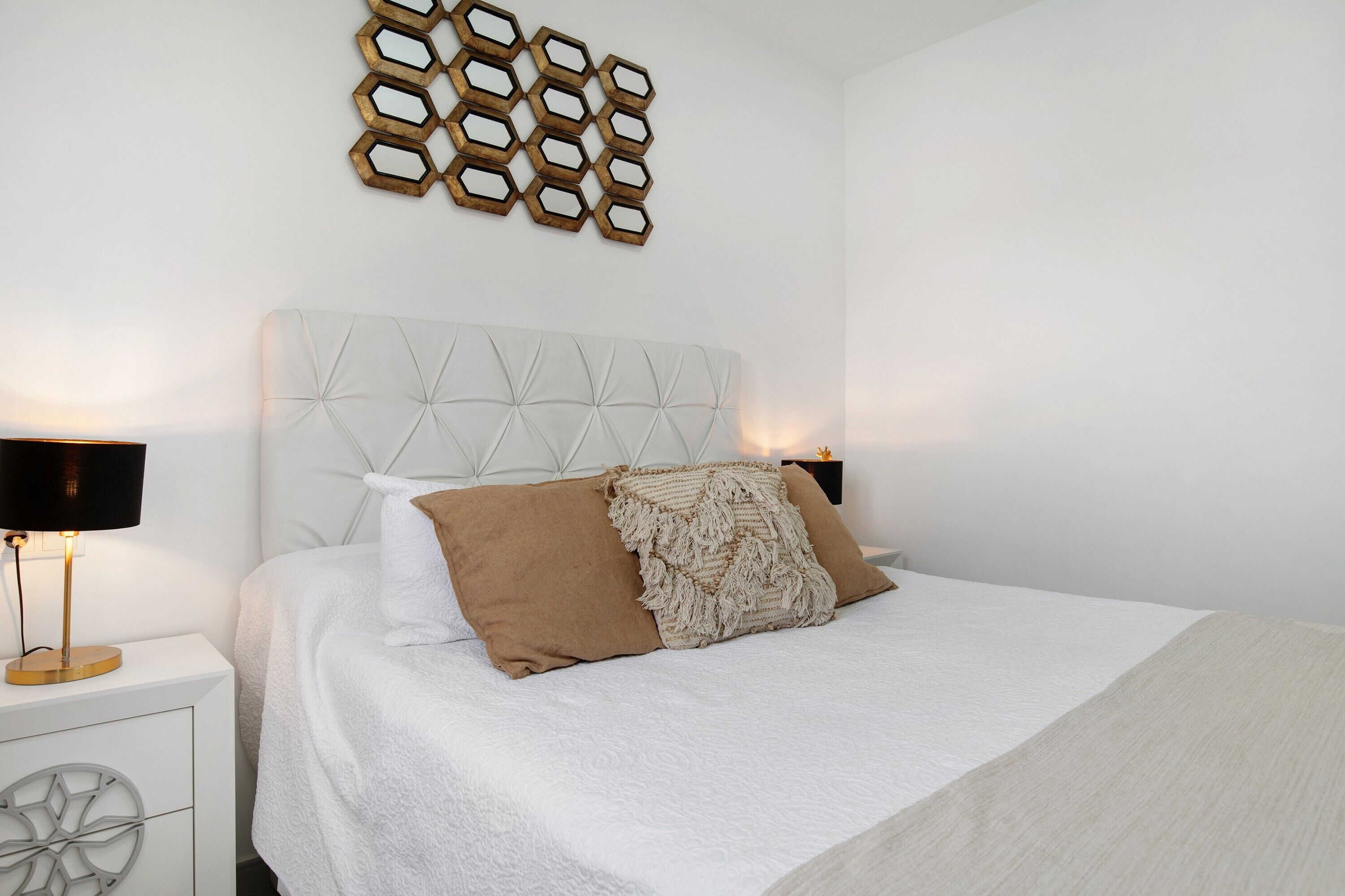 A bedroom with a tufted headboard, two bedside lamps, a decorative wall piece, and a bed with a white and beige bedspread.