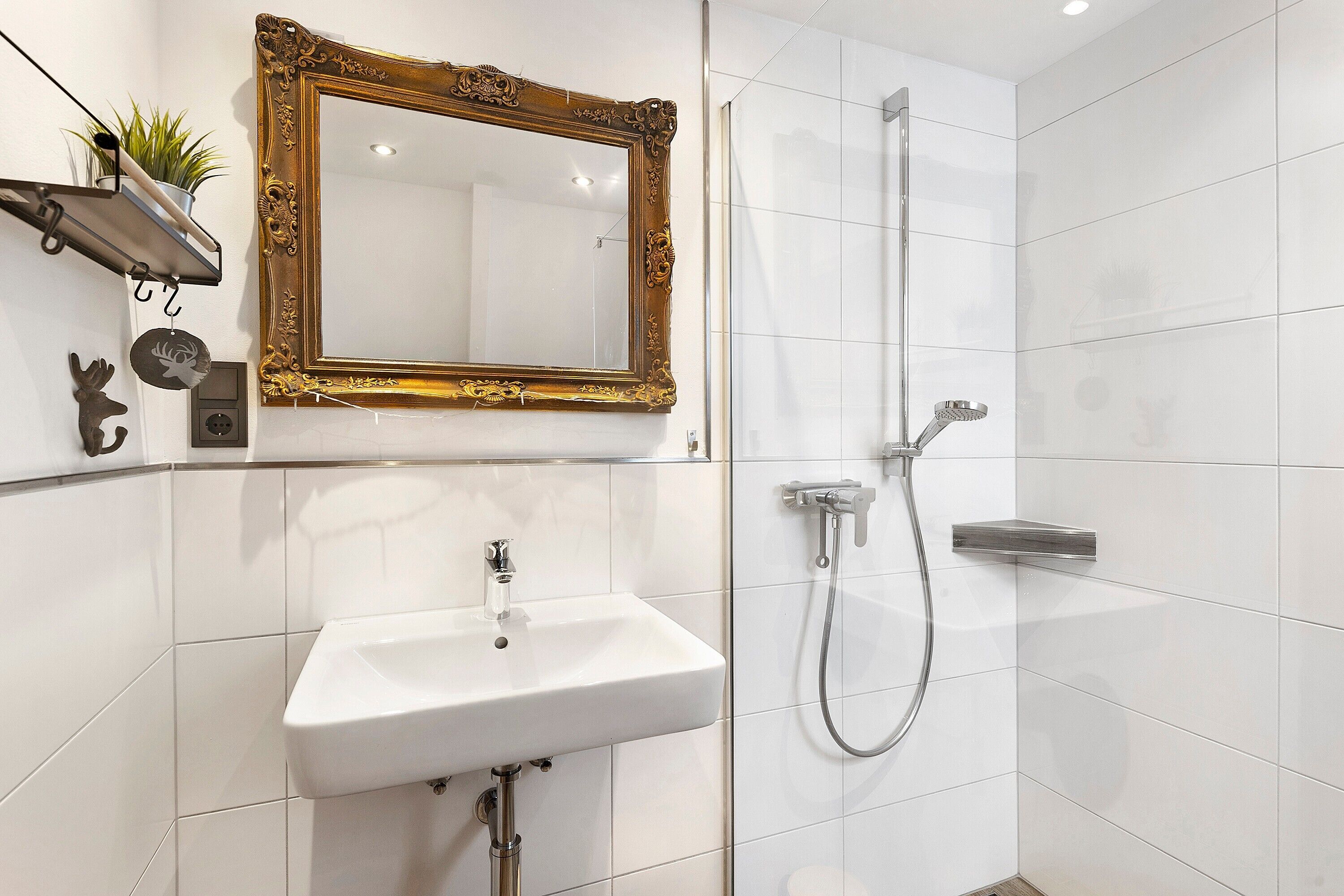 Une salle de bain avec un lavabo blanc, un miroir à cadre doré, une douche et une petite étagère sur laquelle est posée une plante.