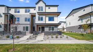 A modern multi-story residential building with a central entrance, a small front yard with a tree, and a clear blue sky. -  New townhome, quick freeway & public trans. access, seasonal pool & pickleball. (American Fork)