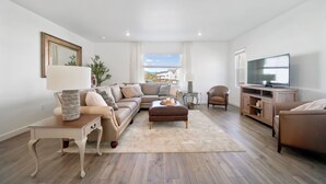 A modern living room with a sectional sofa, a coffee table, a TV stand, and a large window providing a view of the outside. -  New townhome, quick freeway & public trans. access, seasonal pool & pickleball. (American Fork)