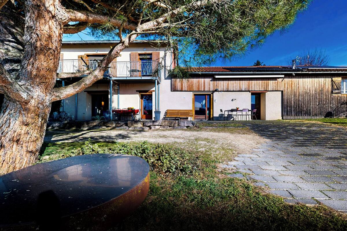 A modern house with a paved courtyard, a dining area with outdoor furniture, and a large tree in the foreground.