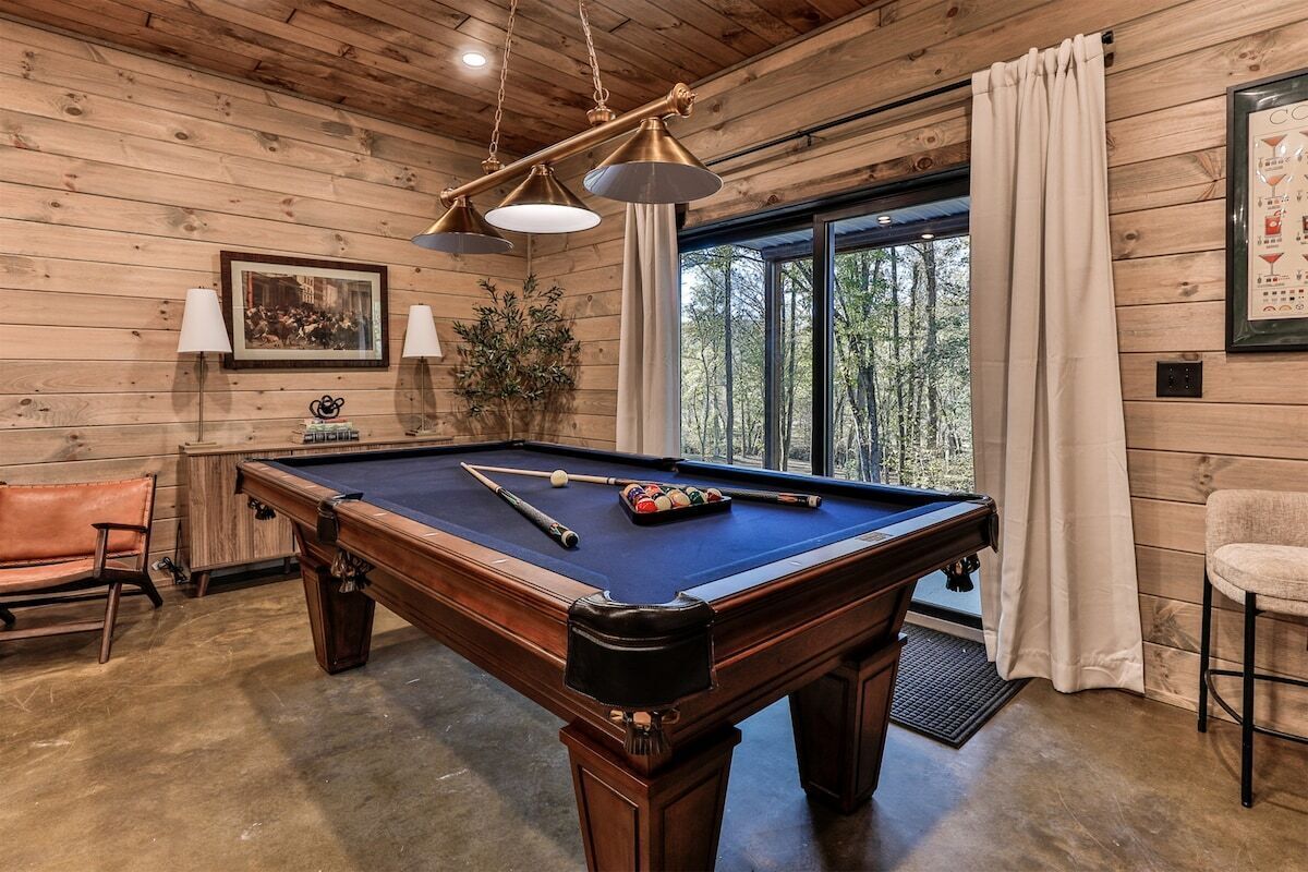 A pool table with cues and balls, a bench, a potted plant, and a window with curtains.