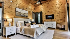 Un dormitorio con techo de madera, cama con ropa de cama blanca, mesita de noche, cómoda y una silla.