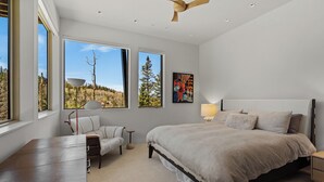 Un dormitorio con una cama grande, un escritorio y dos ventanas que dan a vista de árboles y un cielo despejado.